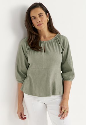 Kobieta z długimi brązowymi włosami, nosząca złote kolczyki koła, zieloną teksturowaną bluzkę z rękawami 3/4 i białe spodnie, stojąca przy białej ścianie.