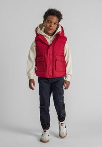 Rote Pufferweste mit Kapuze, ausgestattet mit Fronttaschen, über einem hellen Hoodie getragen, kombiniert mit dunklen Cargo-Hosen und weißen Sneakers.