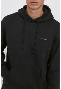 Sudadera con capucha negra hecha de un tejido suave, con un bolsillo tipo canguro. Presenta un pequeño logo en el pecho y cordones ajustables en la capucha.
