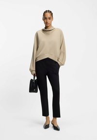 Beiger übergroßer Pullover mit hohem Kragen, kombiniert mit schwarzen zulaufenden Hosen. Schwarze Handtasche und schwarze spitze Ballerinas vervollständigen den Look.