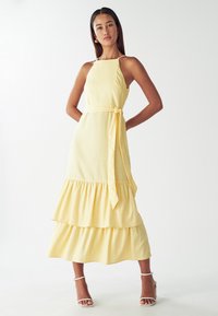 Geel gelaagd jurkje met spaghettibandjes, een aansluitende body, een gestrikte taille en gerimpelde lagen aan de onderkant. Paar witte sandaaltjes met hak.