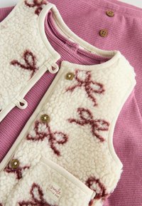 Cremfarbener Fleece-Weste mit braunen Schleifenmustern, beigem Rand und goldenen Knöpfen, schichtweise über einem strukturierten blush-pinken Grund.