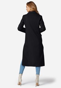 Langer schwarzer Mantel mit strukturiertem Muster, Reverskragen und taillierter Passform, kombiniert mit hellblauen Jeans und cremefarbenen Stiefeletten.