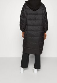 Sort pufferjakke med en lang silhuet, quiltet tekstur og overdimensioneret hætte. Har lynlåse i siderne og hviler over hvide sko.