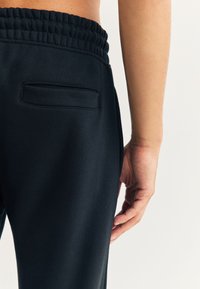Zwarte katoenen joggingbroek met een elastische tailleband, voorzien van een achterzak en een gestructureerde afwerking. De zijaanzicht toont een close-up van de stof.