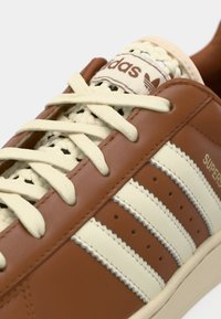 Zapatilla deportiva de cuero marrón con rayas crema, cordones tejidos, detalles en color blanco roto y diseño texturizado. Cuenta con el logo de la marca en la lengüeta.