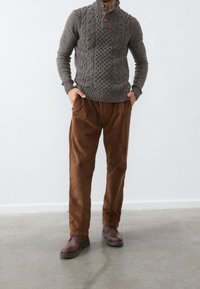 Maglione grigio lavorato a maglia con trecce strutturate, pantaloni di velluto a coste marroni e scarpe marroni con lacci. Il maglione presenta un collo alto e dettagli con bottoni.