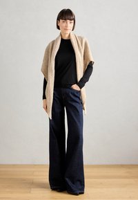 Châle tricoté beige sur un haut à manches longues noir, associé à un jean large bleu foncé. Fond en bois.