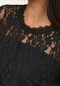 Svart spetsöverdel med blommönster, vågig halsringning och texturerad kropp. Genomskinliga sektioner framhäver intrikata detaljer i hela plagget.
