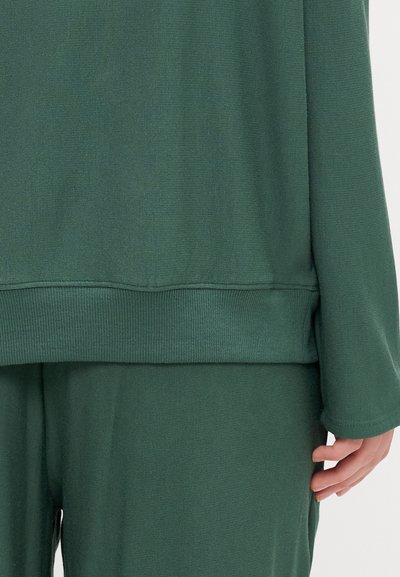 Sweatshirt verde com punhos e bainha canelados, textura macia e um ajuste descontraído. Detalhes de costura visíveis. Combinado com calças verdes a condizer.