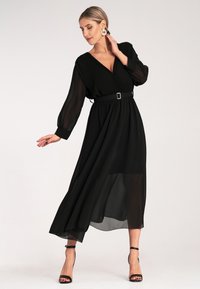 Czarna sukienka z plisowanego szyfonu z głębokim dekoltem w kształcie litery V, długimi rękawami i paskiem w talii z prostokątną klamrą; szpilki na kostkę.