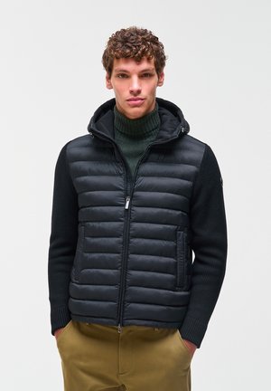 Giovane uomo con capelli ricci indossa una giacca imbottita nera con cappuccio, dolcevita verde scuro e pantaloni marroni, in piedi davanti a uno sfondo semplice.