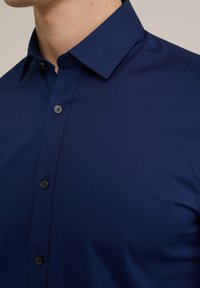Cămașă bleumarin cu nasturi, realizată dintr-un material de bumbac neted, având un guler structurat și detalii subtile cu nasturi pe partea din față.