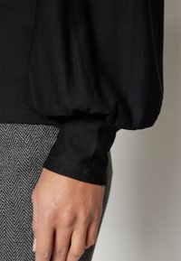 Black, soft fabric with voluminous sleeves, paired with herringbone-patterned gray material at the bottom, showcasing a fitted cuff detail.