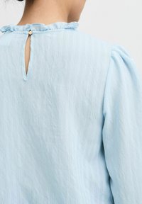 Top azzurro chiaro con scollatura arricciata, apertura a chiave sul retro e realizzato in un tessuto testurizzato con sottili linee verticali.