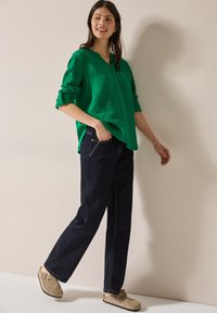 Camicia button-up verde con texture e maniche rimboccate, abbinata a jeans blu scuro e scarpe slip-on beige. Sfondo semplice.