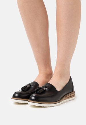 Mocassins en cuir noir avec des pampilles portés pieds nus, dotés de semelles en caoutchouc blanc et de détails en marron superposés au-dessus de la semelle.