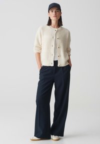 Cremfarbener Strickcardigan mit strukturierten Mustern, sechs Knöpfen, kombiniert mit einer dunkelblauen weit geschnittenen Hose und einer blauen Mütze. Sneaker runden das Outfit ab.