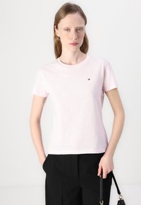 Tricou roz deschis cu mânecă scurtă, realizat dintr-un material moale din bumbac. Prezintă un mic logo brodat pe piept. Este asortat cu pantaloni negri.