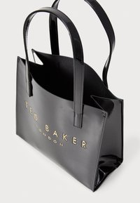 Sac fourre-tout noir en cuir verni avec deux poignées, arborant une marque dorée à l'avant et une doublure intérieure douce. Design simple et structuré.