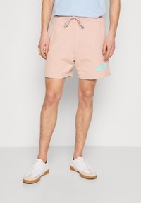 Shorts en mélange de coton rose avec une taille élastique, un cordon de serrage, et un détail de logo bleu sur la jambe droite. Portés avec des baskets blanches.