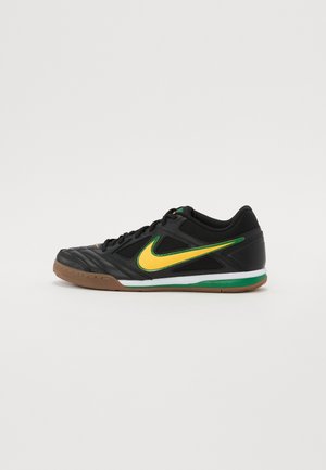 Zapatilla negra de fútbol sala con el logo Nike Swoosh en amarillo y verde, suela de goma y diseño de perfil bajo sobre un fondo blanco liso.