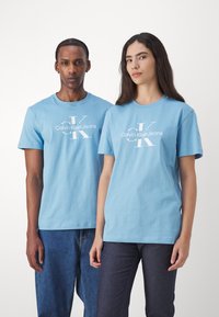 Gaiši zila kokvilnas T-krekls ar centrālo balto logo "Calvin Klein Jeans", ar apaļu kakla izgriezumu un īsām piedurknēm.