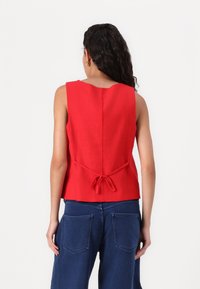 Red sleeveless top with a round neckline, featuring a tied waist detail and a smooth texture. Paired with dark blue wide-leg jeans.