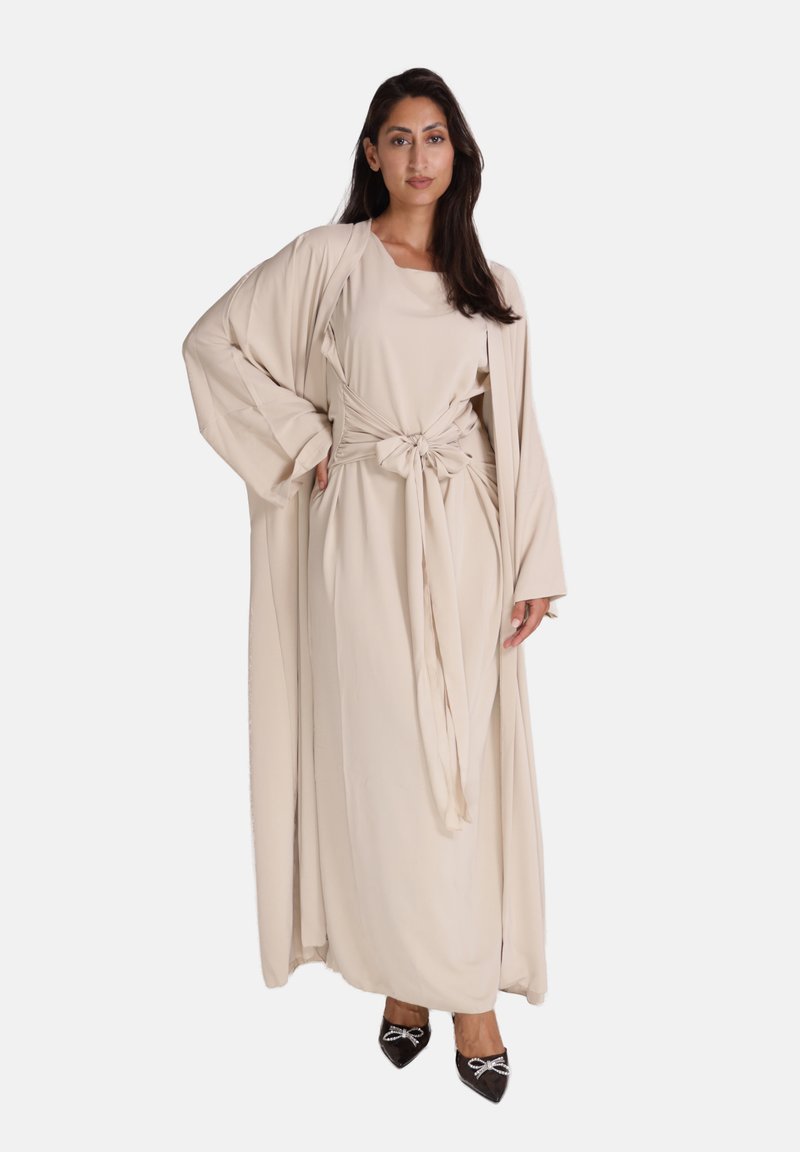 Beige lange jurk met een gestrikte taille en brede mouwen, gelaagd met een bijpassende open voorjak en gecombineerd met zwarte puntige schoenen.