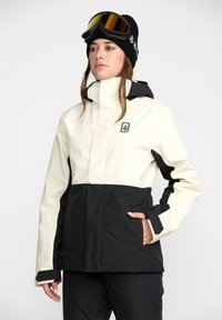 Ski jakke med en hvid øverste sektion og sort nederste sektion. Har hætte, lynlåslommer og logo på brystet. Glat tekstur.