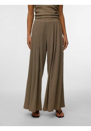 Donna che indossa pantaloni marroni a vita alta e a gamba larga e sandali neri con cinturini, in piedi su uno sfondo chiaro e semplice.