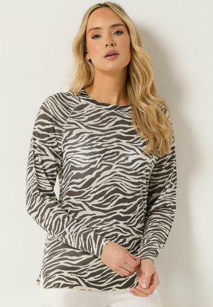 Frau mit langen blonden Haaren, die ein langärmliges, zebragestreiftes Hemd trägt und vor einer schlichten hellen Wand posiert.