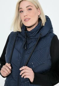 Kvinna med blont hår justerar snörena på en marinblå mönstrad pufferväst som bärs över en svart långärmad topp mot en vit bakgrund.