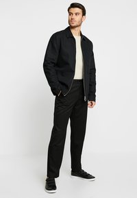 Schwarze Zip-Up-Jacke über einem hellen Pullover, kombiniert mit einer schwarzen Hose und schwarzen Sneakern. Die Jacke hat einen Kragen und seitliche Taschen.