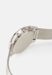 Silver stainless steel watch with a mesh band, round case, and two side buttons. Smooth textured back with engraved details.