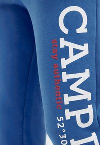 Blauer Stoff mit weißen, fetten Buchstaben "CAMP" und rotem, vertikalem Text "stay authentic" neben weißen Koordinaten "52°30'".