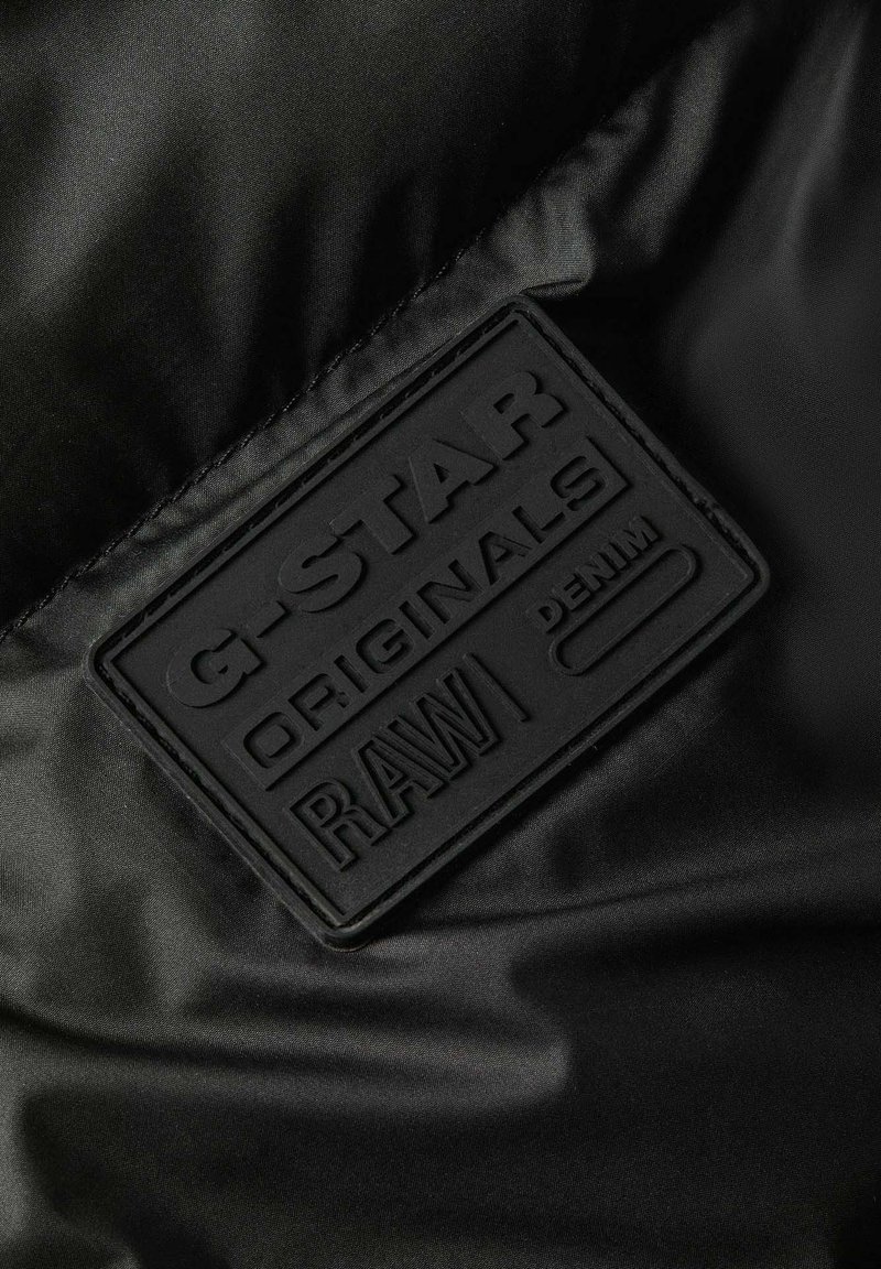 Schwarzes Stoffetikett mit eingeprägtem Text "G-STAR ORIGINALS RAW DENIM" auf einem glatten schwarzen Textil-Hintergrund.
