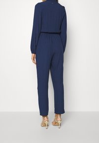 Navy blue jumpsuit features long sleeves, elastic waistband, and tapered legs with a textured fabric. Worn with gold strappy heels.
