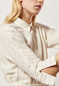 Biała koszula w złote pionowe paski, z kołnierzykiem i podwiniętymi rękawami, które są przytrzymywane na miejscu przez guziki.