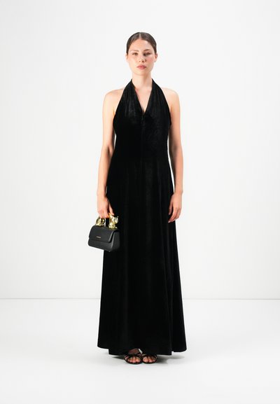 Woman in a long black halter dress holding a small black handbag with gold animal-shaped handles, standing against a plain white background.