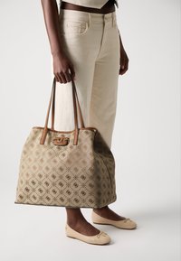 Osoba w kremowych spodniach i beżowych balerinach trzyma dużą torbę w kolorze jasnobrązowym z wzorem, z brązowymi skórzanymi uchwytami i emblematem logo.