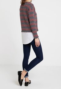 Haut à manches longues à motifs multicolores superposé à une chemise blanche, associé à un jean skinny foncé et des mocassins noirs.