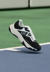 Schwarz-weißer EA7 Emporio Armani Sneaker, der über einer blau-grünen Tennisplatzoberfläche schwebt.