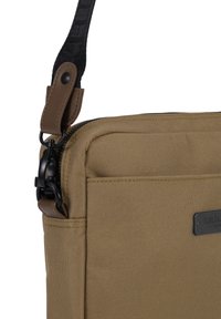 Borsa a spalla rettangolare in tela color sabbia con hardware nero, chiusura con zip e una tracolla regolabile con dettaglio in pelle e etichetta con logo.