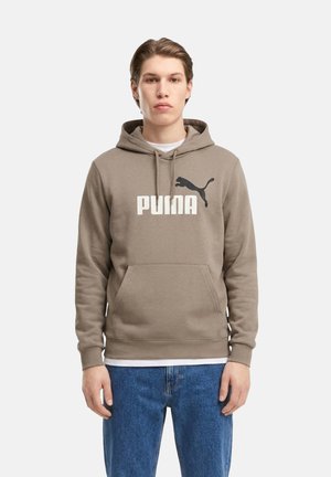 Giovane uomo che indossa una felpa con cappuccio taupe Puma con logo davanti e jeans blu, in piedi con espressione neutra rivolto in avanti.