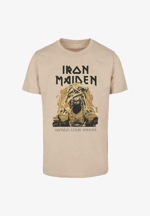Béžové bavlnené tričko s grafikou kostry v reťaziach, s nápisom "Iron Maiden" nad a "World Tour 1984-85" pod ním v čiernej farbe.