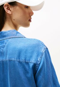 Camicia di jeans blu chiaro con colletto, caratterizzata da una texture uniforme e cuciture visibili. La parte posteriore della camicia è mostrata, rivelando i dettagli del design.