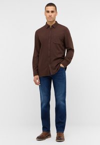 Brązowa koszula z długim rękawem, z kieszonką z przodu, zapinana na guziki, w połączeniu z niebieskimi dżinsami i brązowymi skórzanymi butami, wszystko na jasnoszarym tle.