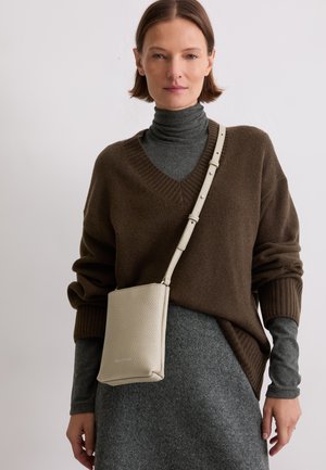 Beige leren crossbodytas met een gestructureerd oppervlak en een minimalistisch ontwerp, voorzien van een lange verstelbare riem en een vierkante vorm, gedragen bij een donkere outfit.
