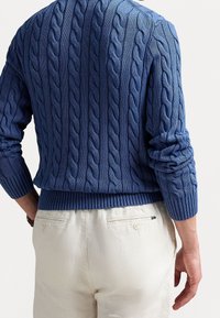 Mies, jolla on sininen kaapelineulottu villapaita, jossa on resorit hihansuissa ja helmassa, yhdistettynä luonnonvalkoisiin housuihin, joissa on nappikiinnitteiset takataskut.